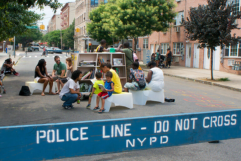 A view of people reading on an open street. There's a blue barricade in the foreground that reads "POLICE LINE - DO NOT CROSS NYPD"