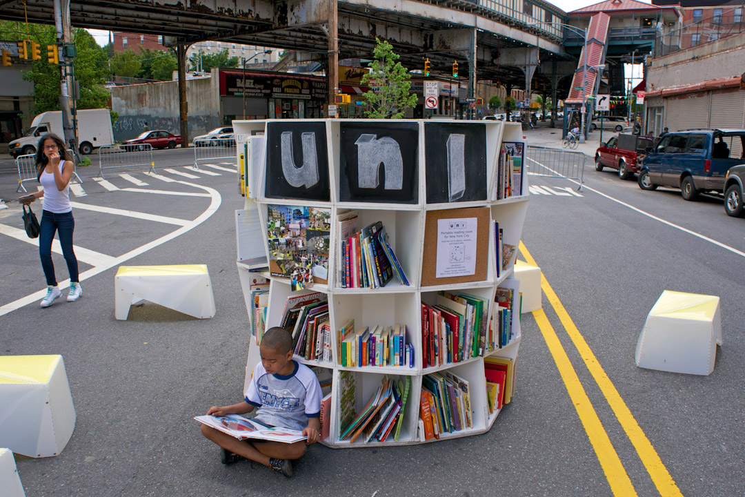 A boy reading a book on the street, leaning on a white bookshelf tower.
