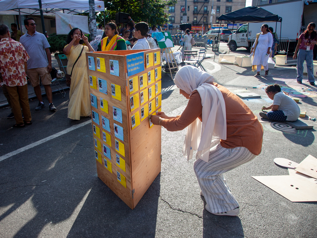 A woman putting up a yellow card with polaroid picture on a wooden board for part of the community engagement activity in a street in NYC.