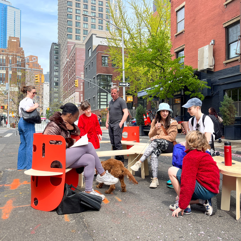 A photograph of people sitting and talking on a red and wooden bench in the street 