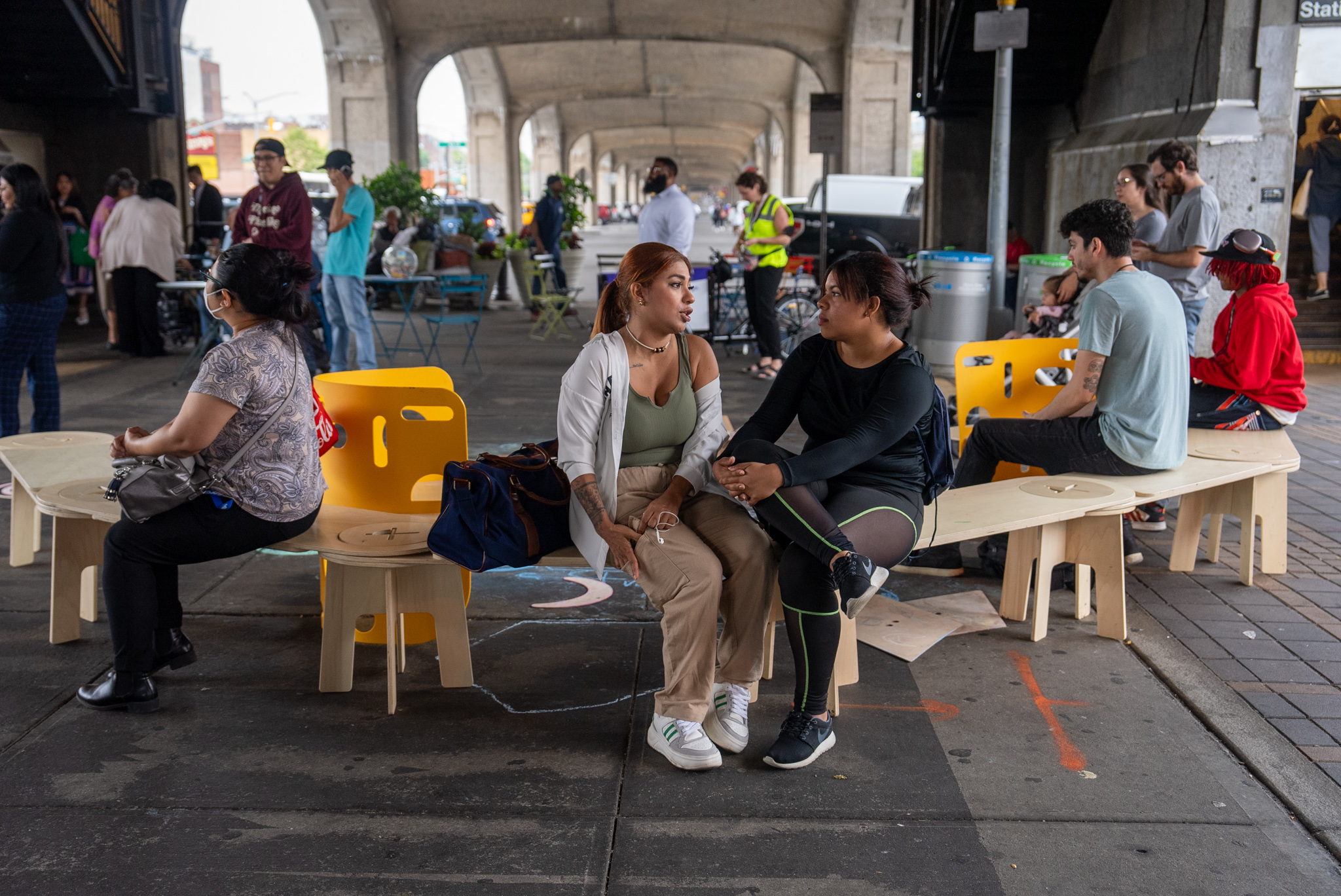 a photograph of people sitting on a wooden and plastic bench under a bridge 