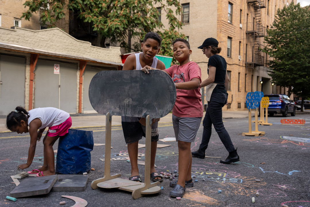 Two children looking at the camera and posing on an open street in NYC. Another child is chalking on the ground, on the left of the two children.