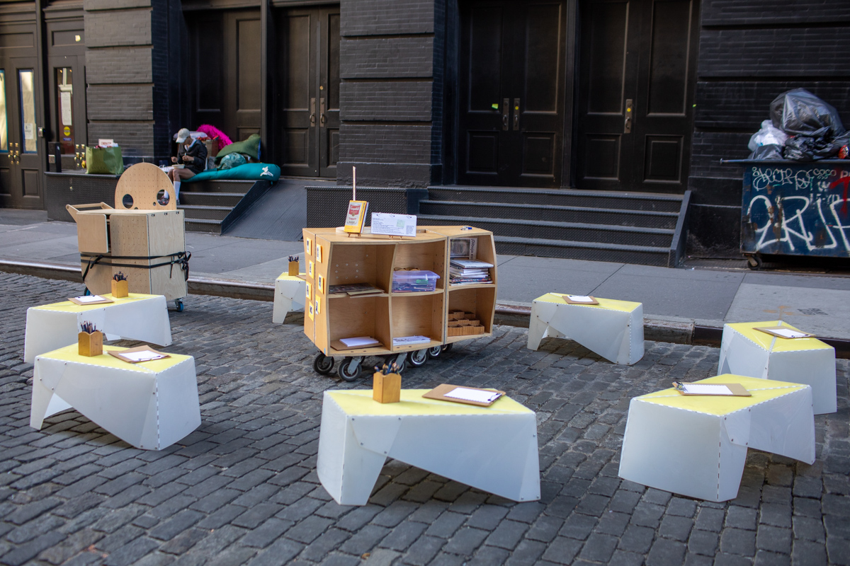 A photograph of portable wooden cart with drawing materials and plastic benches surrounding it in a street in NYC.