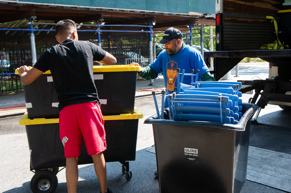 Two men moving boxes and containers, with one container showing metal chairs; on a street in NYC.
