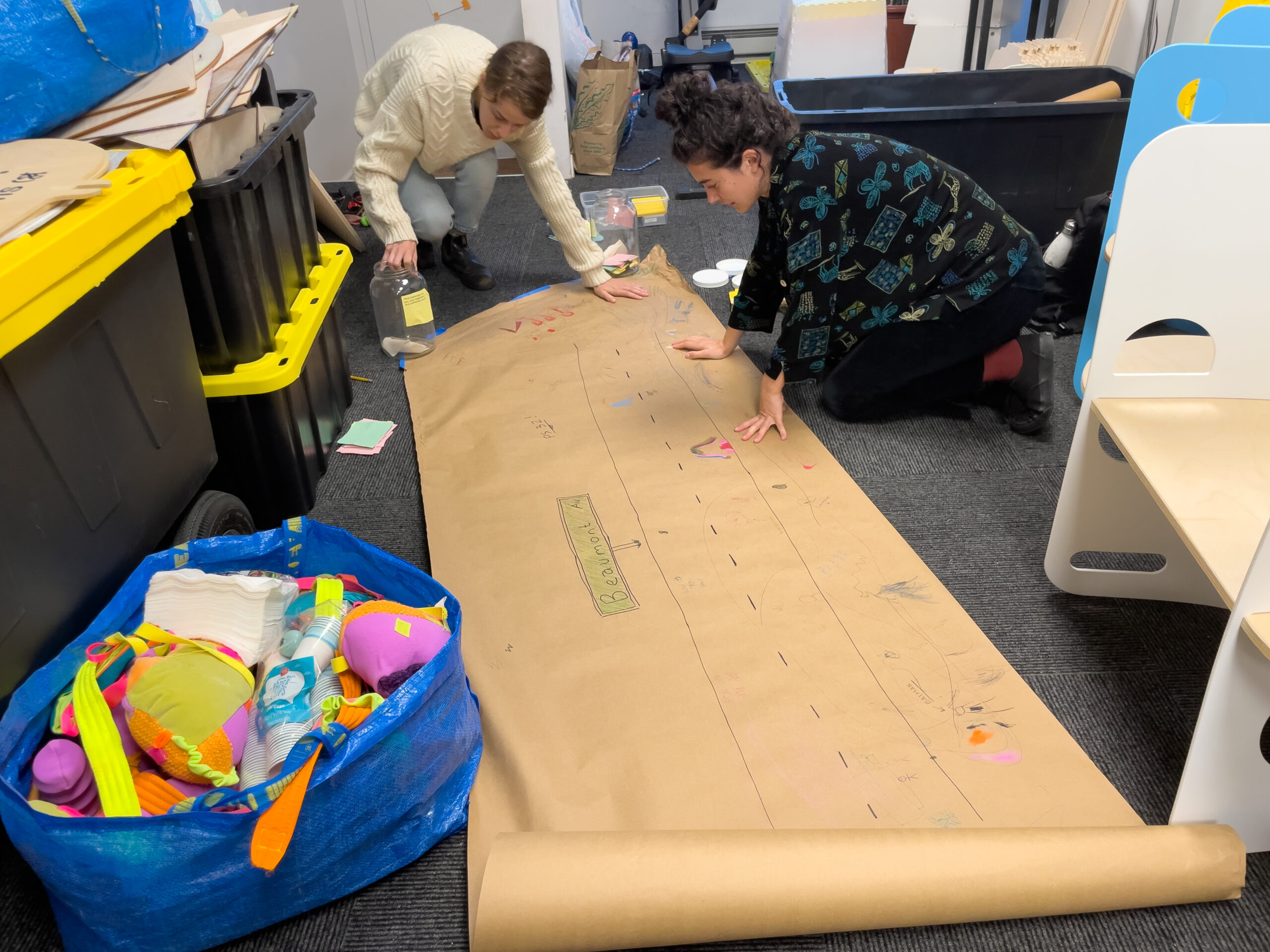 Two women working on the floor with a roll of craft paper and drawings on it.