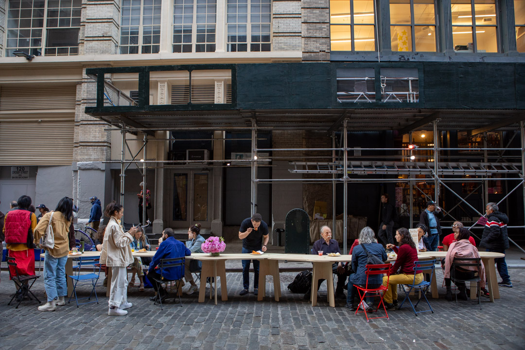 A group of people learning how to embroider on one big table on the street of NYC.