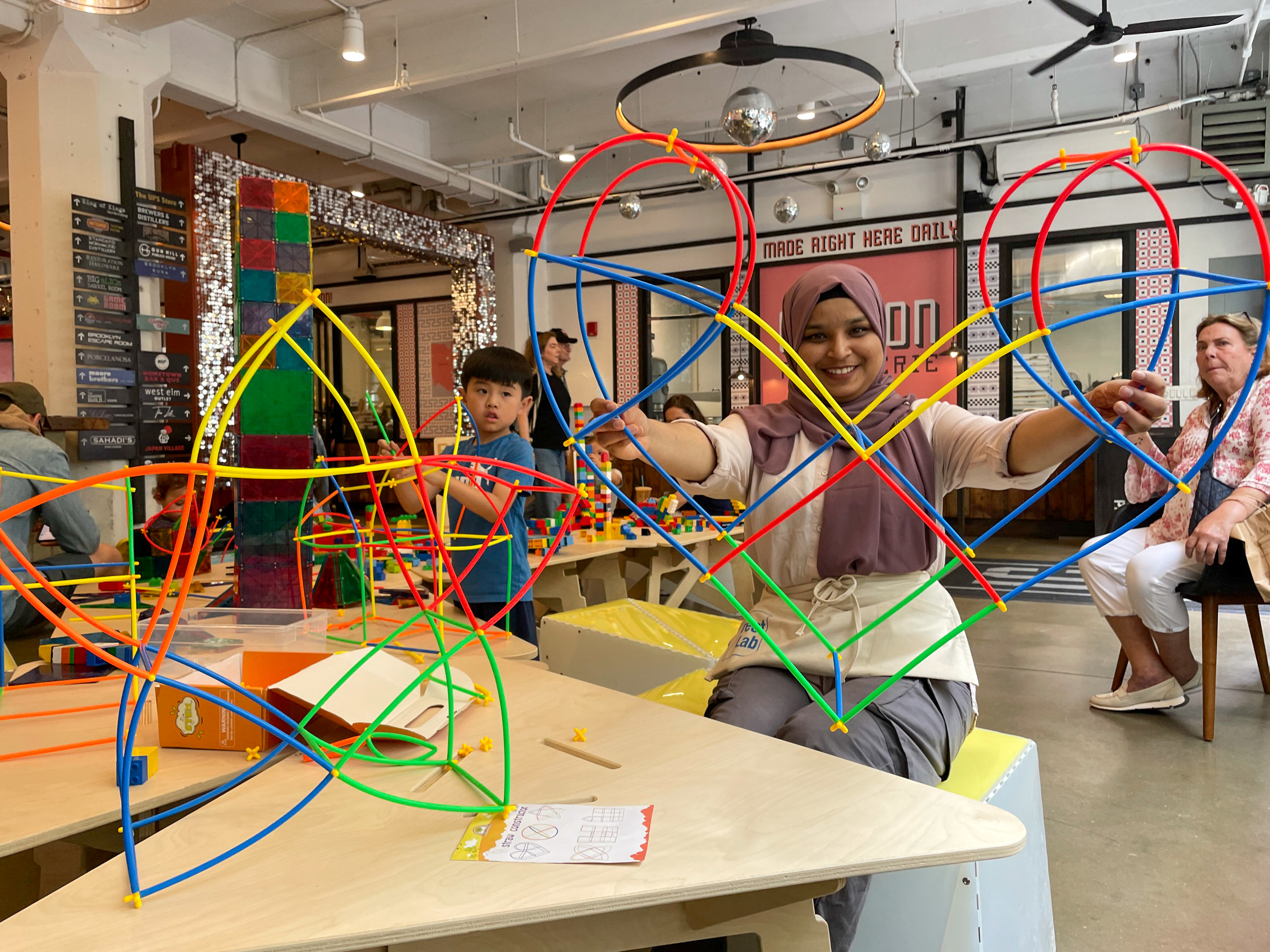 A woman holding a hard-shaped structure made with colorful straw toys in an indoor market.