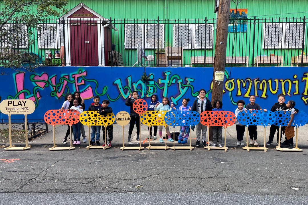 A photo of children in a straight line facing the camera, with colorful play obstacles in front and school building in the back.