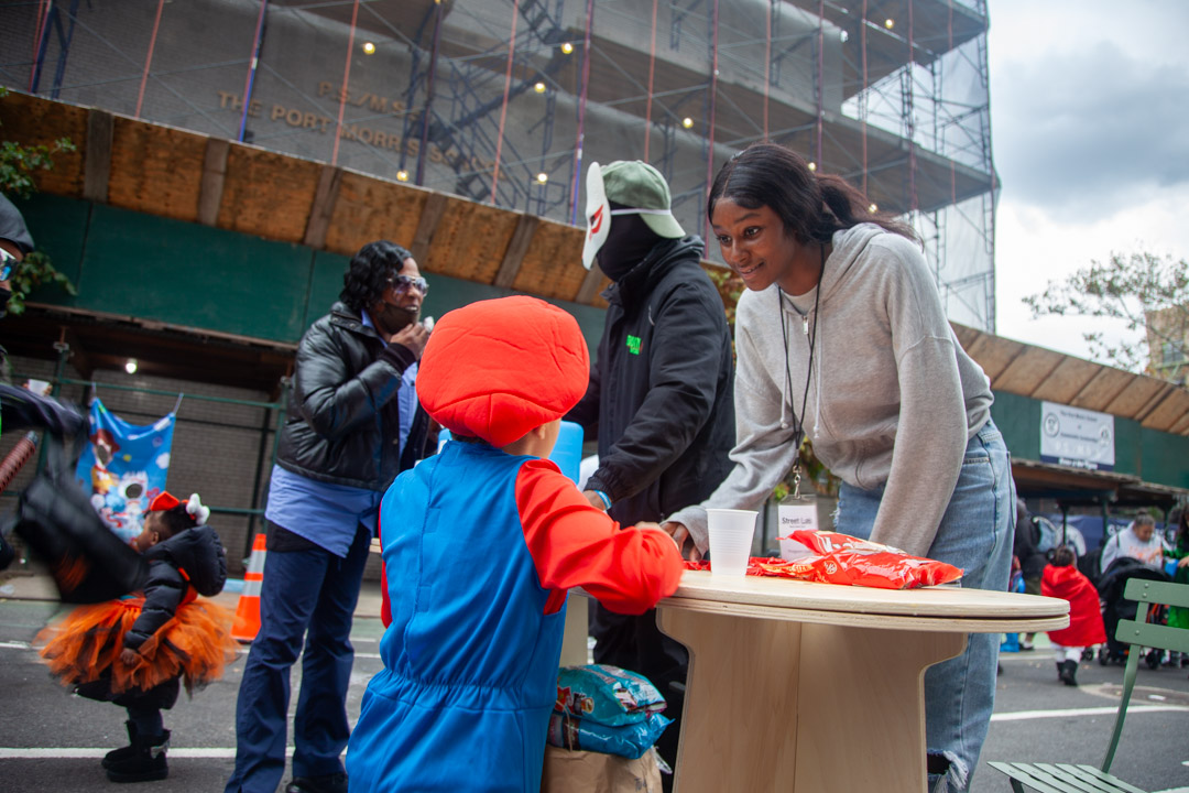 A woman wearing a gray hoodie and a man wearing a mask giving out candy in an open street during Halloween.