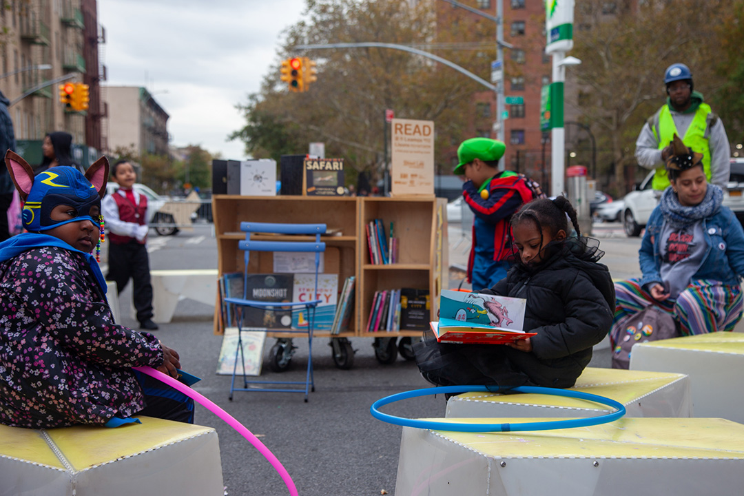 Some children sitting on plastic benches surrounding a wooden cart with books in an open street in NYC.
