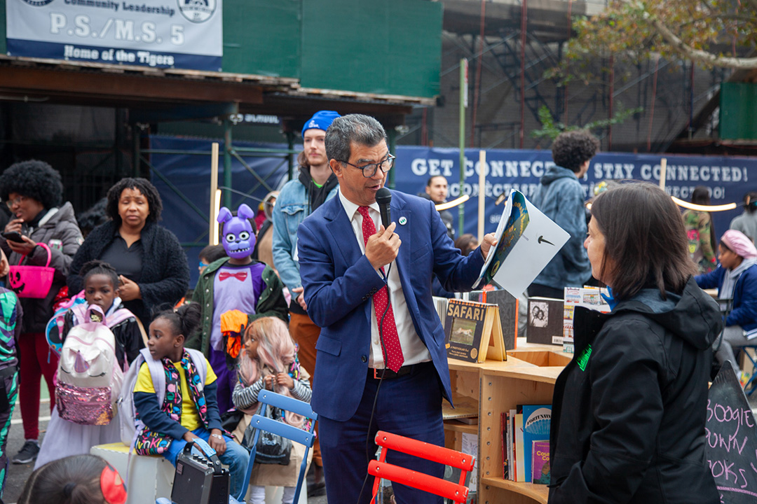 A man in a suit reading a story to a group of children in an open street.