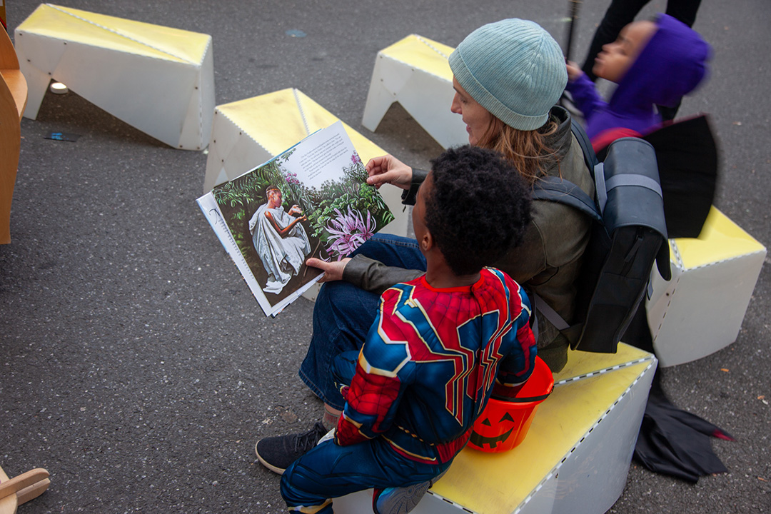 A woman reading a picture book to a boy on a plastic bench in a street.