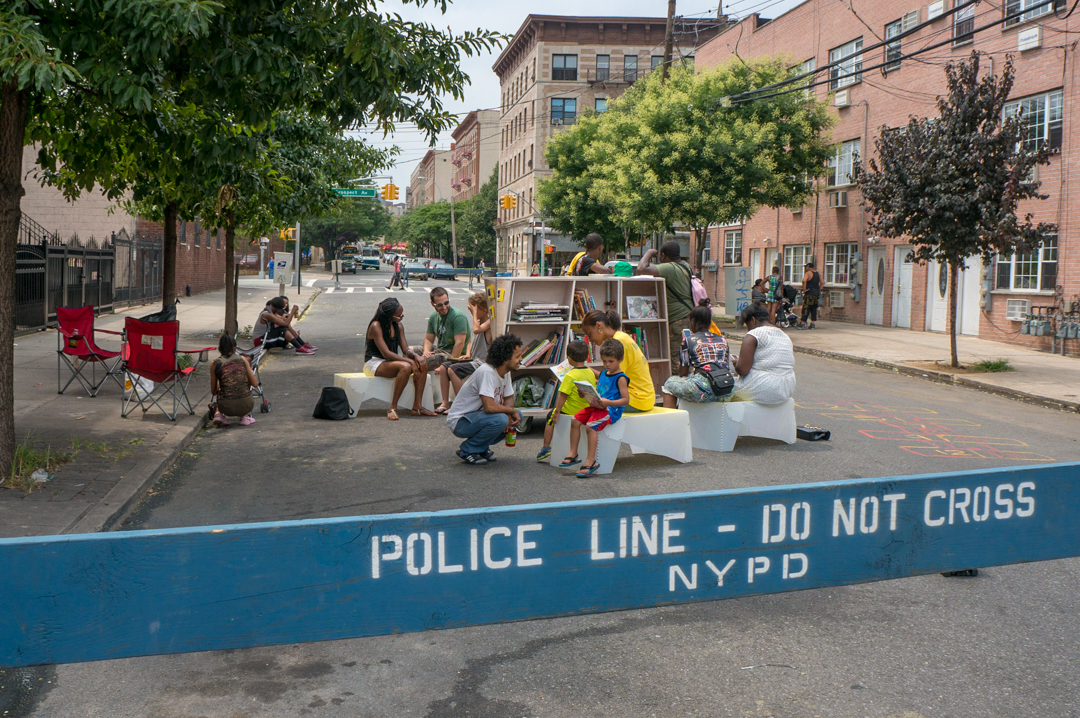 A view of the street with children and adults reading. There is a navy bar that reads "Police Line - Don Not Cross NYPD" in white text.