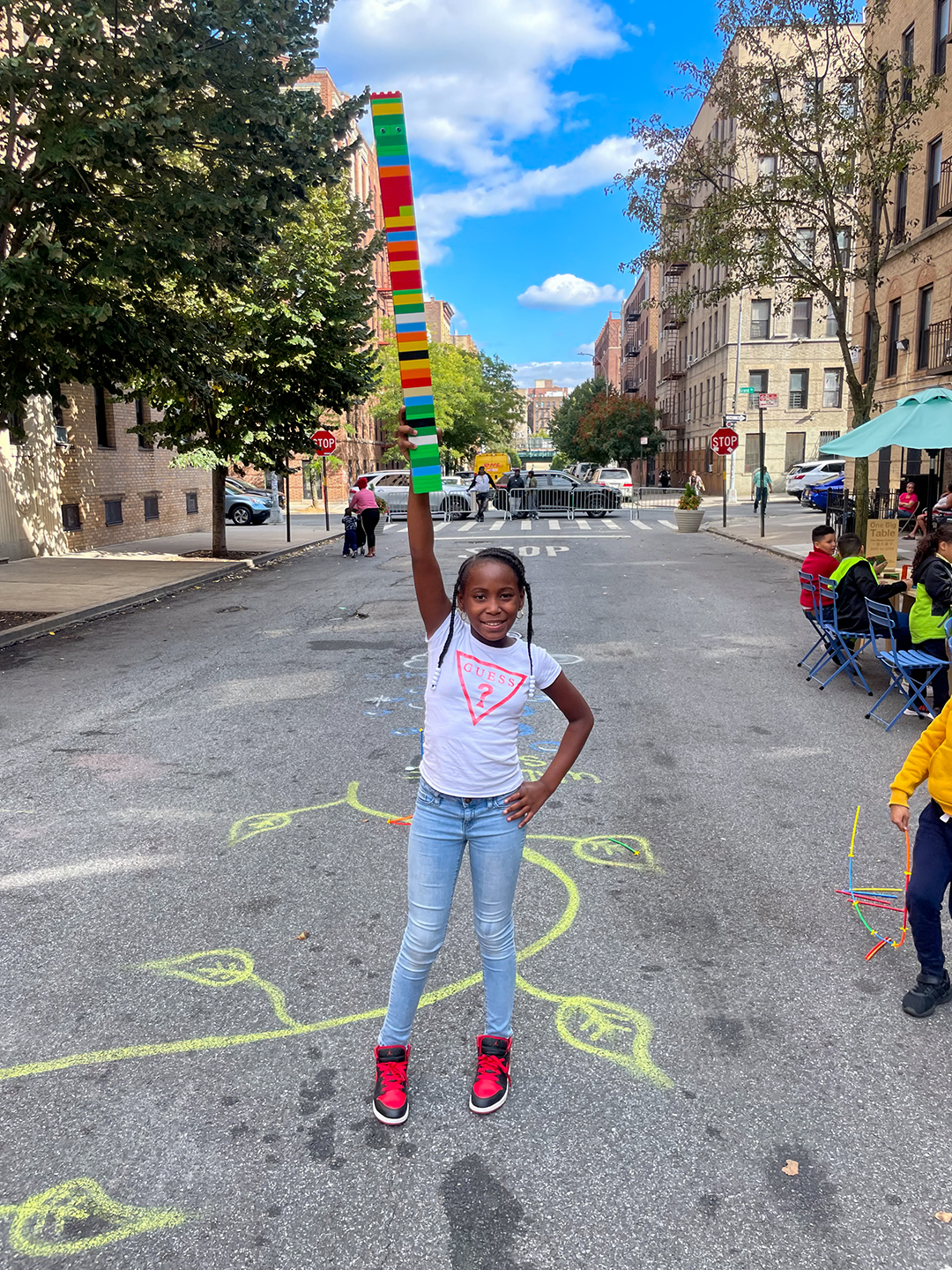 A view of a smiling girl holding up a tall lego tower with one hand on a street of NYC.