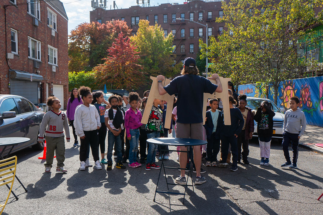 A teacher explaining how to assemble a table to elementary school students on a street in NYC.