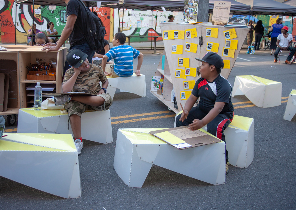 A photo of children in a straight line facing the camera, with colorful play obstacles in front and school building in the back.