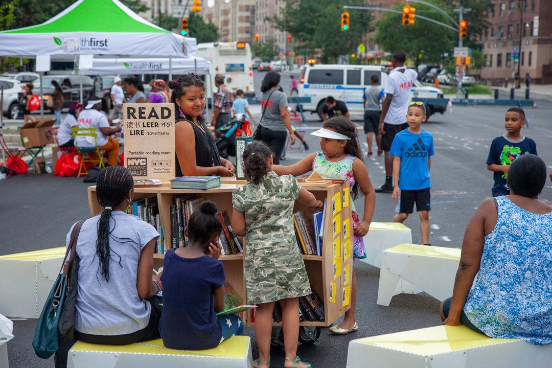 Children and adults gathered around a wooden cart with books and reading, on the street.