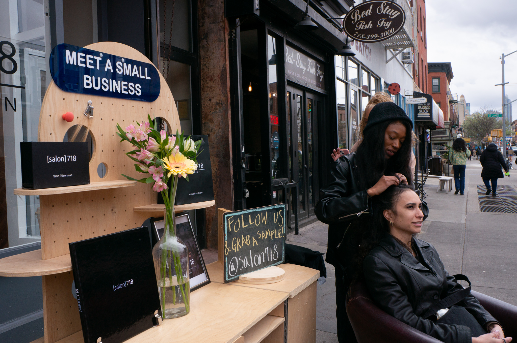 A hair stylist demonstrating a braiding session on the street in front of the storefront.