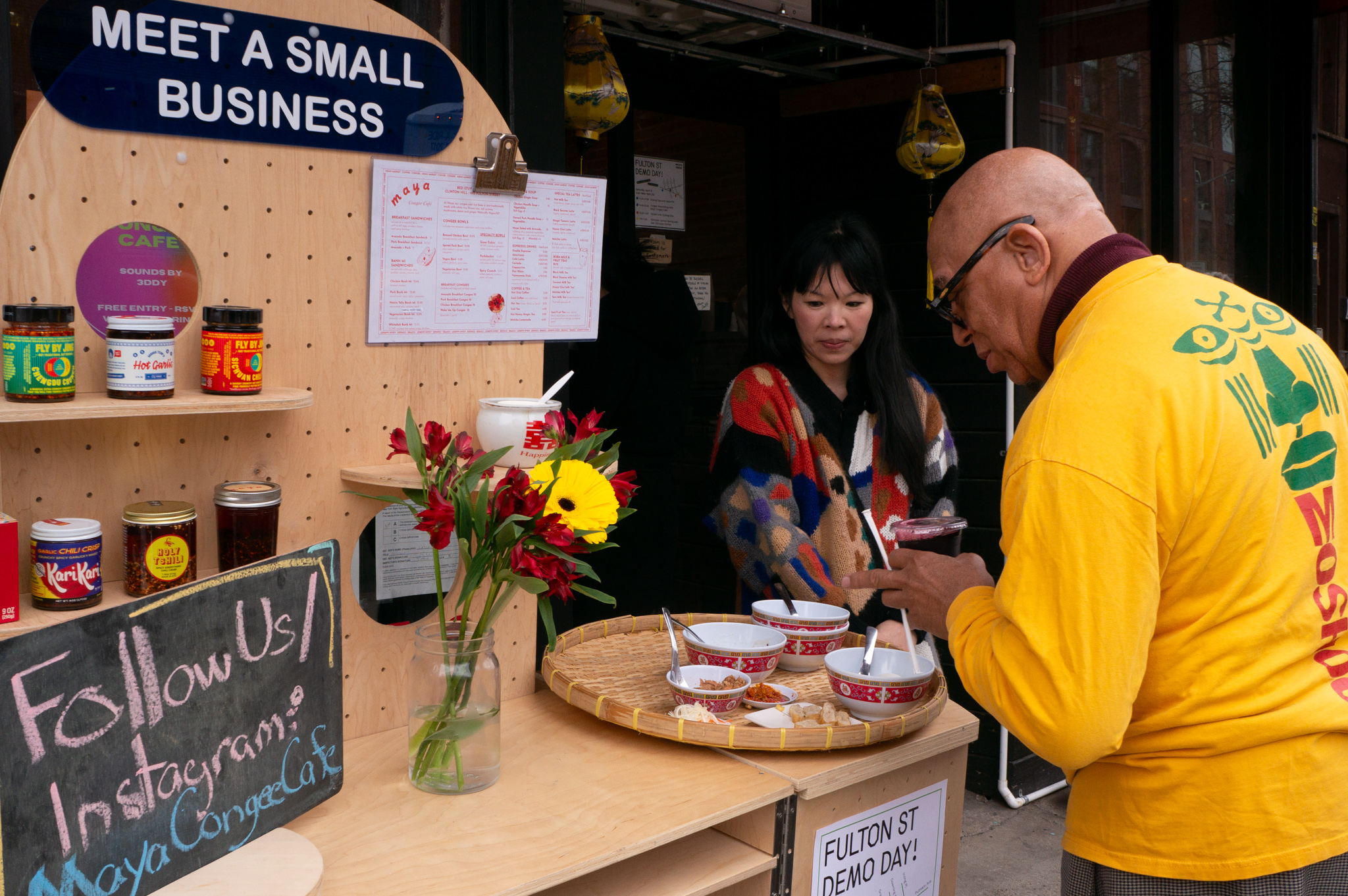 A man looking at a wooden cart on the street with a merchant presenting the food she sells in her restaurant.