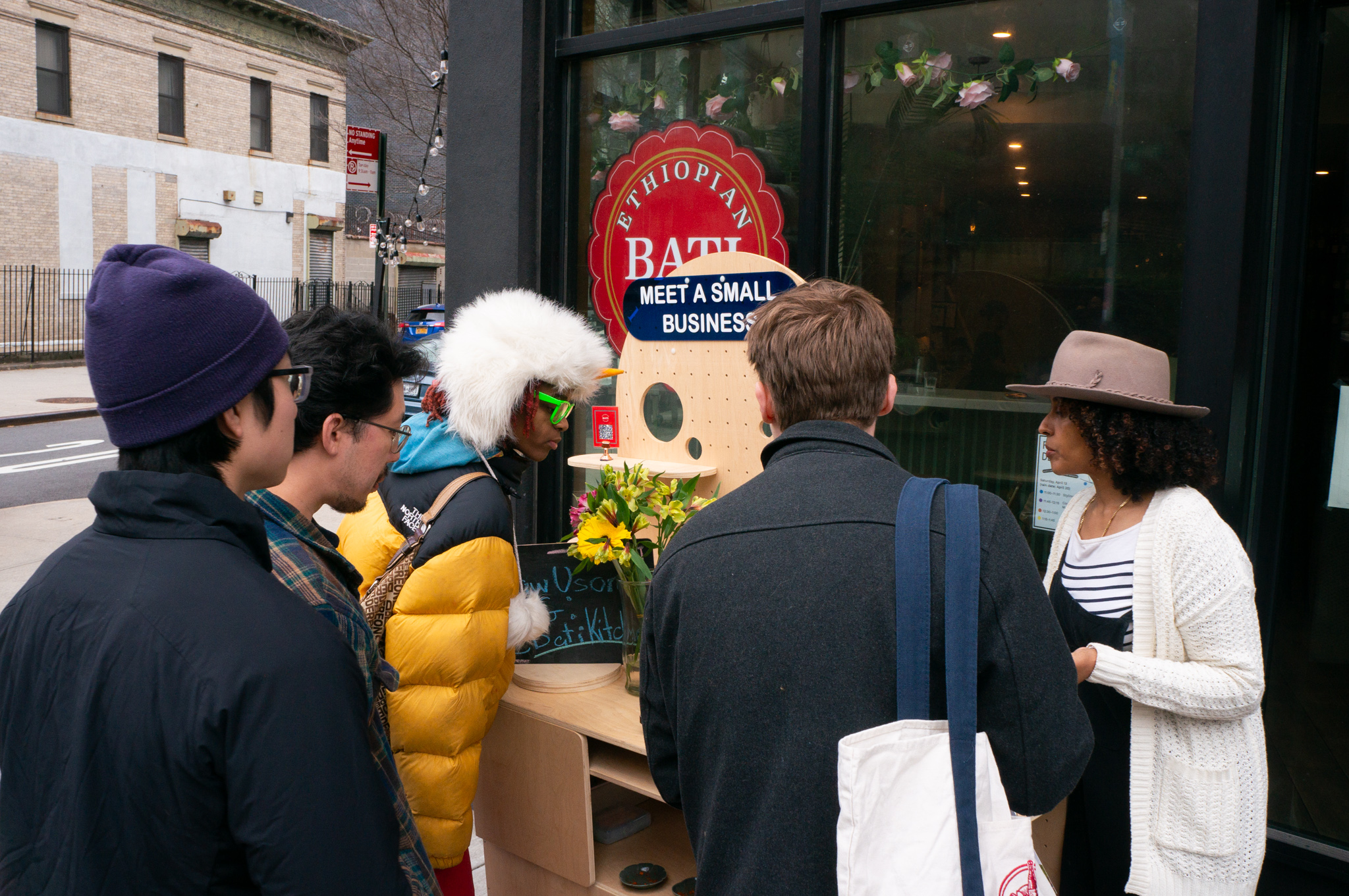 People gathered around a wooden cart in front of the restaurant and the owner demonstrating the things they make in the restaurant.