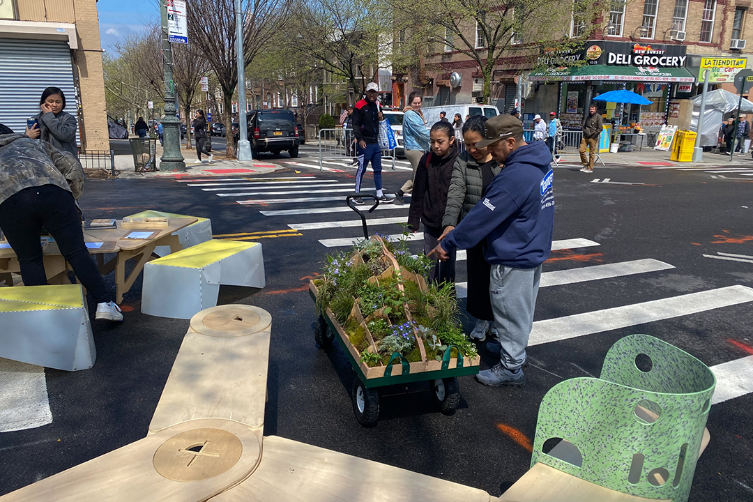 People looking at a wooden cart full of plants on the street.