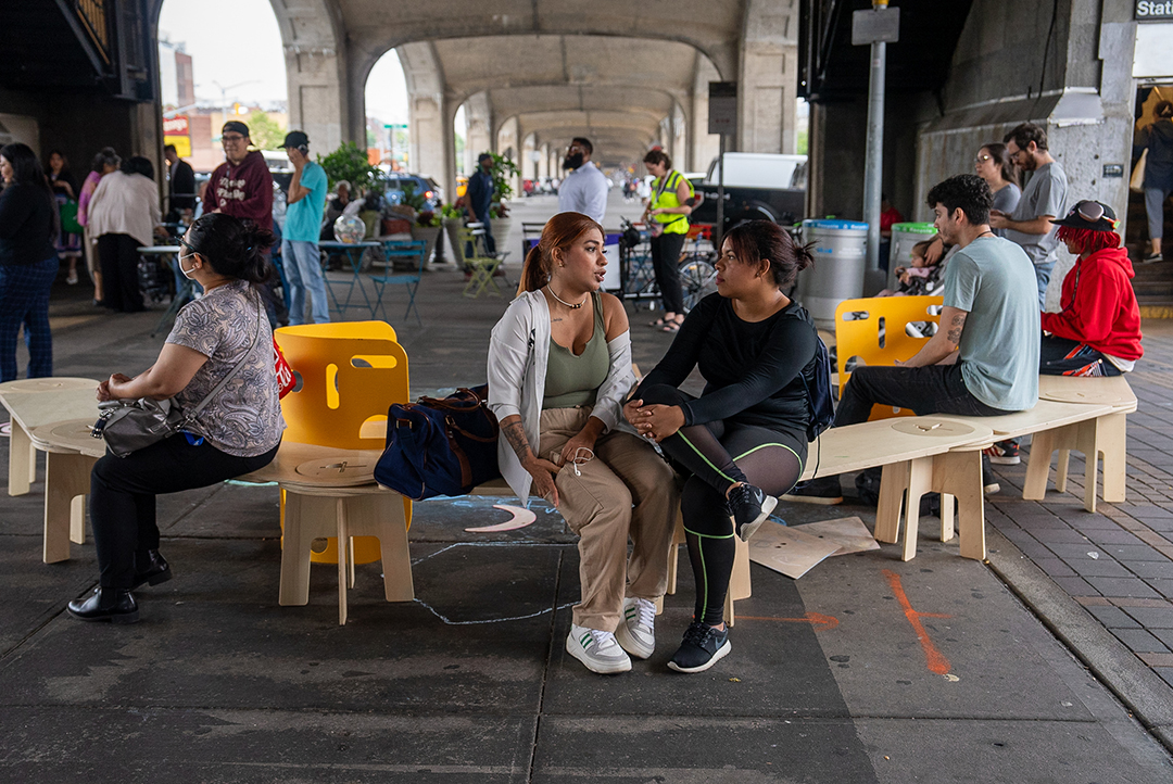 a photograph of people sitting on a wooden and plastic bench under a bridge