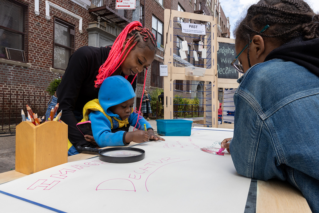 Mom with a child and another older child writing things on the paper on a wooden table on the street.