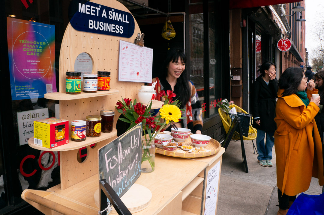 A shop owner standing next to the wooden cart on the street, in front of her storefront.
