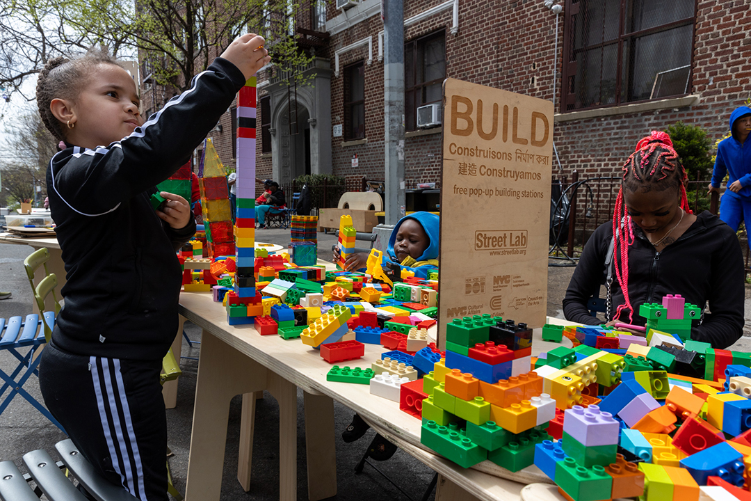 Children and adult playing with lego blocks on a wooden table on the street.