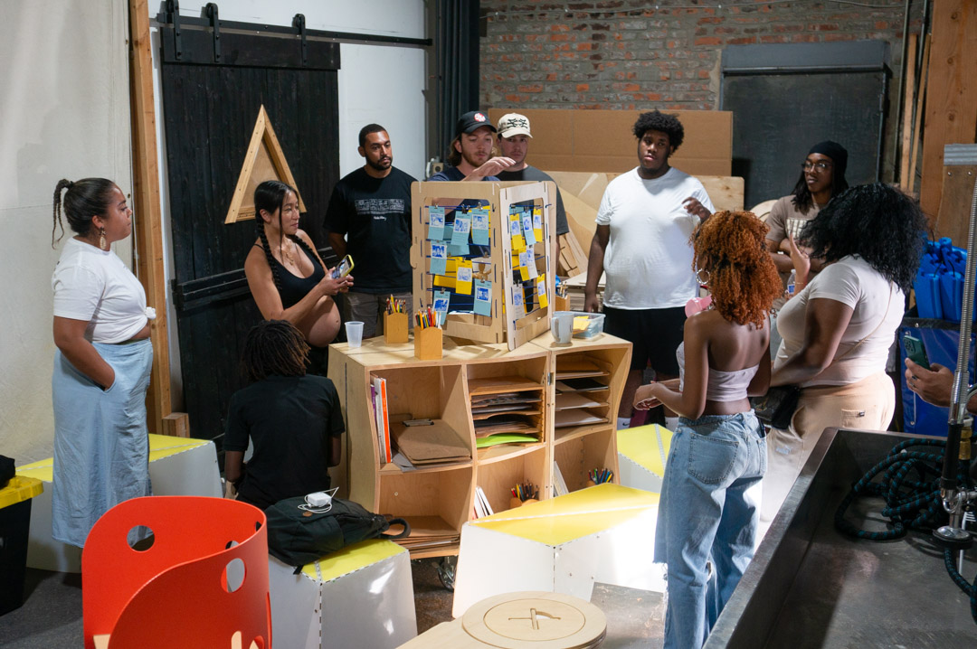 2024-08-13-175007_streetlab-office-warehouse A group of people surrounding a wooden structure, inside a warehouse.