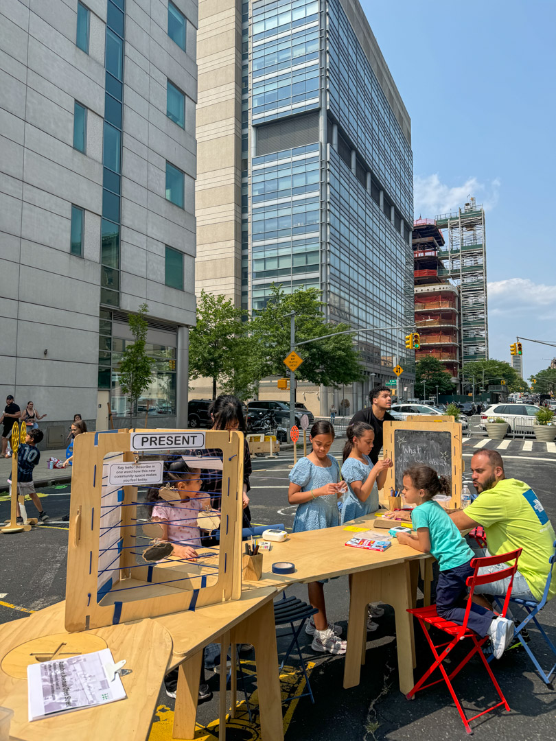 2025-06-11-145250_audubonave_165st-166st Adults and children writing things on the table on a street.