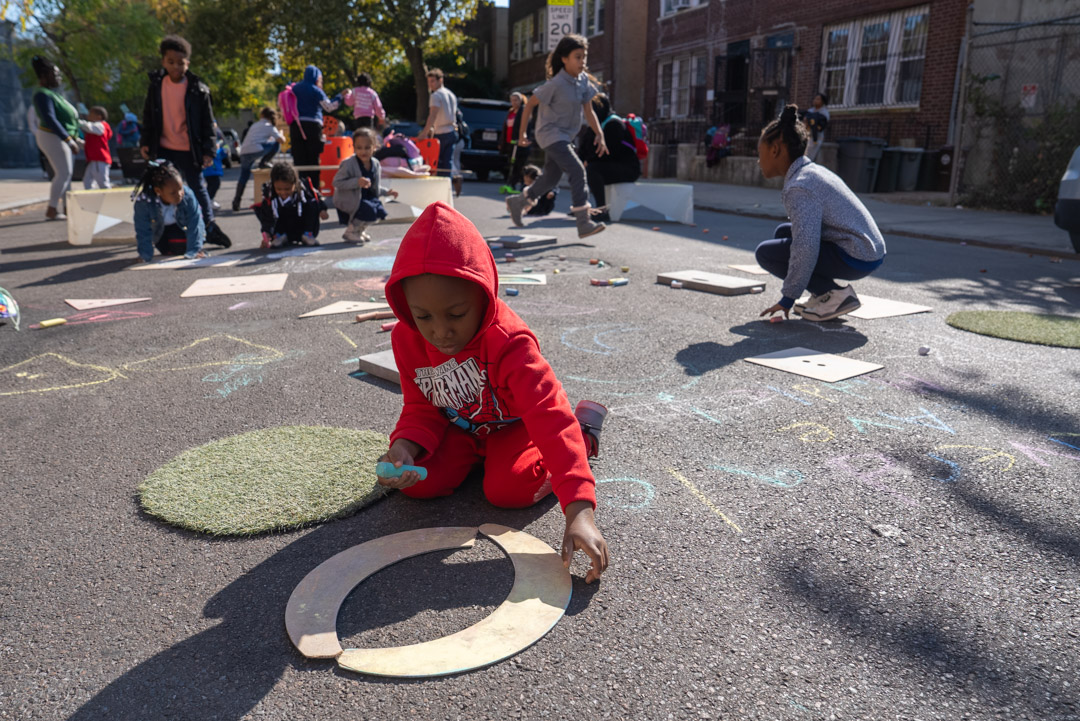 07_2024-10-11-133727_straussst_riverdaleave-newportst_1080px Mom with a child and another older child writing things on the paper on a wooden table on the street.